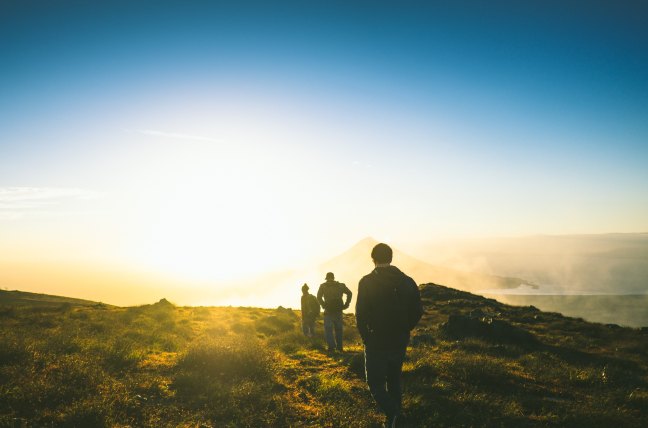 Three men in a single file line on a hill. They are walking away from the camera into a sunset.