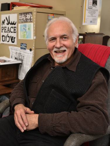 Man with white hair sitting in a chair and smiling at the camera. He is wearing a black vest over a brown shirt.