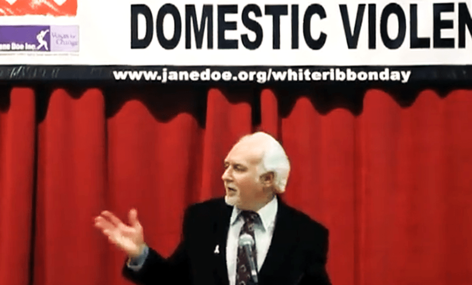 A man with white hair speaking into a microphone in front of a red curtain below a banner that says janedoe.org/whiteribbonday